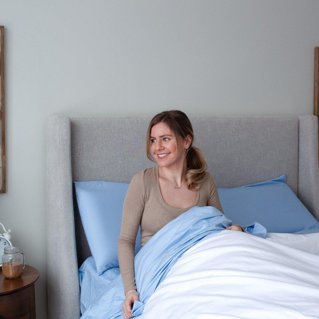 blonde girl sitting up in bed with special edition bahama blue bedface sheet set and white duvet cover against a grey headboard
