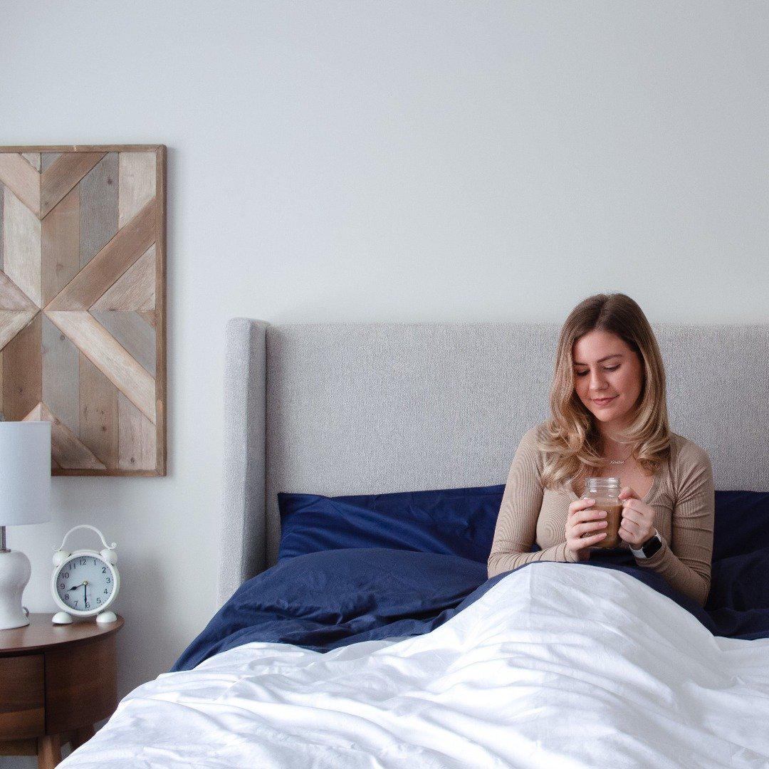 Woman sitting with back against grey fabric headboard starring down at her mason jar mug of coffee tucked into Bedface Nighttine Navy bedding with white weighted blanket on top, folded down