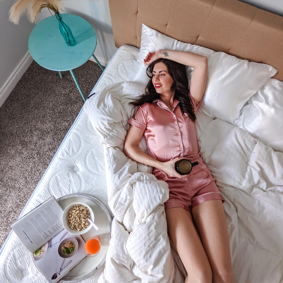 Dark haired woman lying across Haven Mattress in pink satin pajamas holding a coffee with a tray holding cereal and recipe book, with white weighted blanket folded down to show the embossed Haven logo