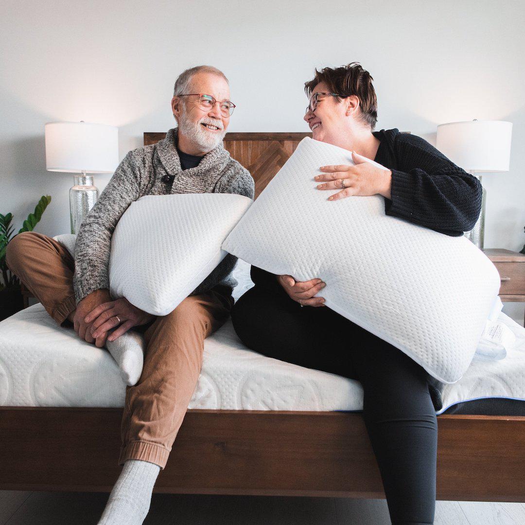 Man and woman both holding Bedface VitaGel 4 in 1 pillows looking at each other smiling while sitting on top of a Haven Mattress