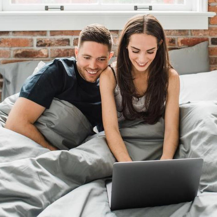 Man and woman cozied up in Haven Mattress bed-in-box bed, tucked into Storm Grey Bedface sheets and duvet cover. The couple is both looking at a grey laptop while the woman is typing