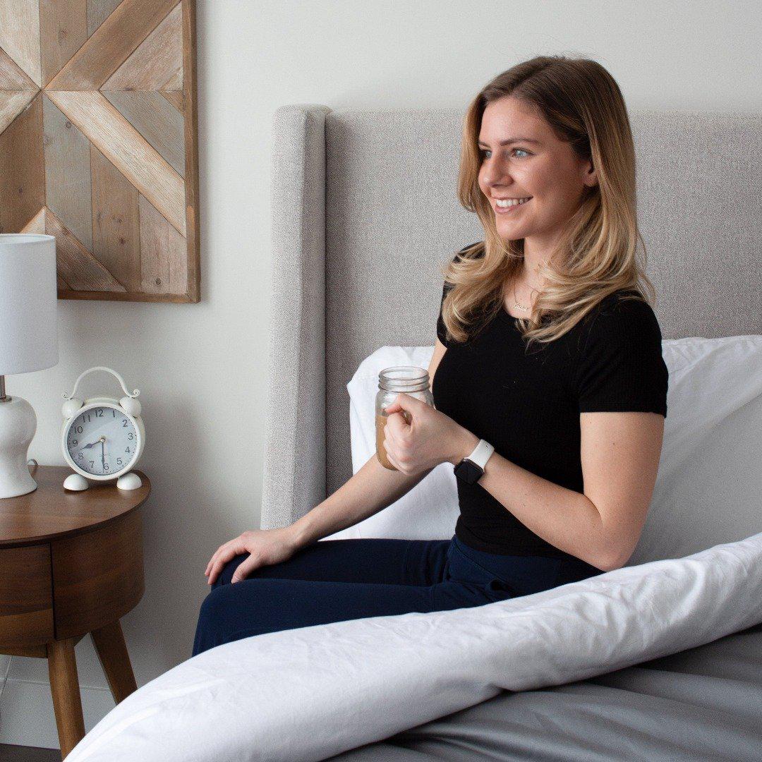 woman sitting upright wearing navy blue outfit holding a mason jar mug with coffee on a Haven Mattress with Starlight White sheet set with Grey weighted blanket turned down