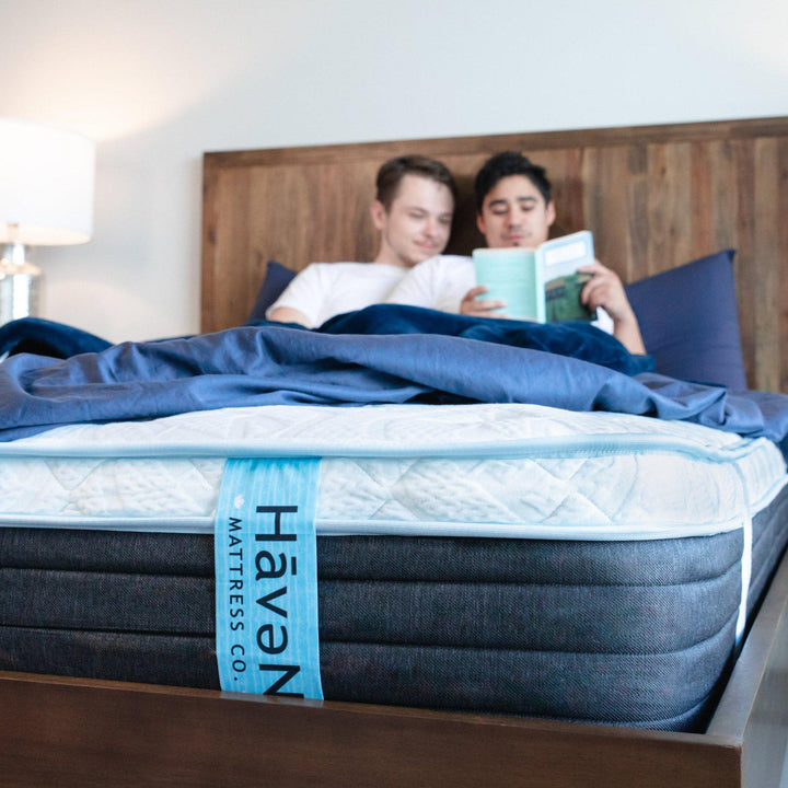 couple laying in bed reading a book together tucked into Nighttime Navy Bedface Sheets. Bottom sheet is folded up to show the Haven Mattress tag down front