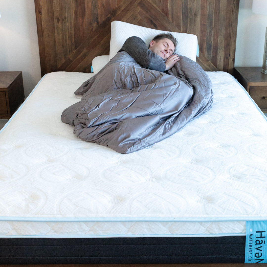 Man curled in Storm Grey weighted blanket showing the embossed Haven Mattress logo with Haven tag down foot of bed