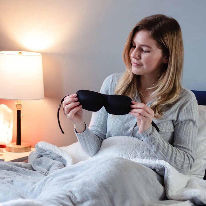woman sitting upright in a Grey Hush Weighted blanket holding a pair of 40 winks blackout sleep mask in black