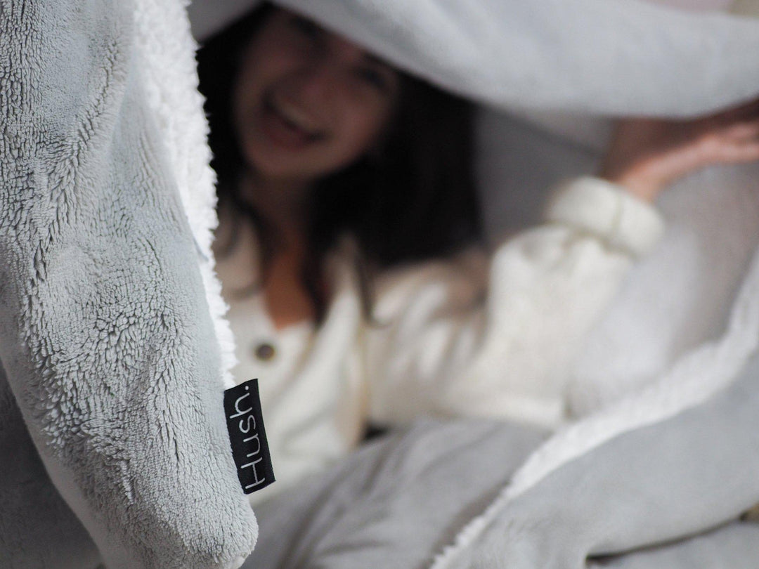 Woman sitting upright wearing a white sweater blown out surrounded by a Hush weighted throw blanket in grey with white fleece liner and a black Hush label tag
