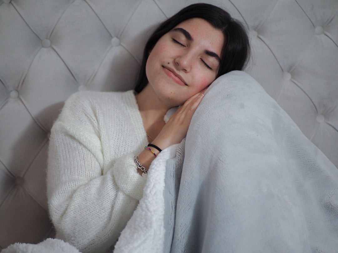 Woman cozied up to a HUSH Weighted Grey Throw with eyes closed with back against a grey fabric headboard