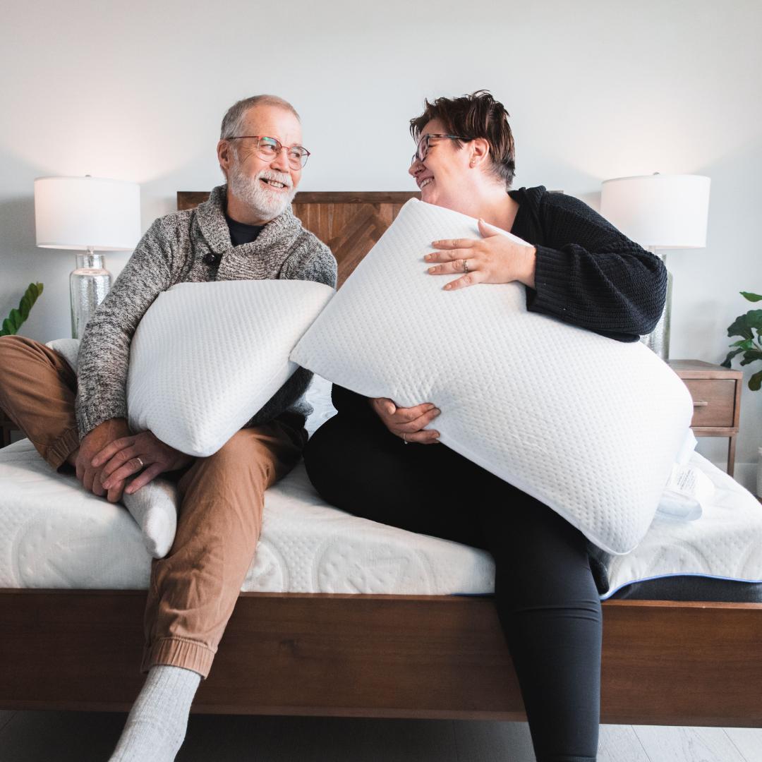 Man and woman both holding Bedface VitaGel 4 in 1 pillows looking at each other smiling while sitting on top of a Haven Mattress