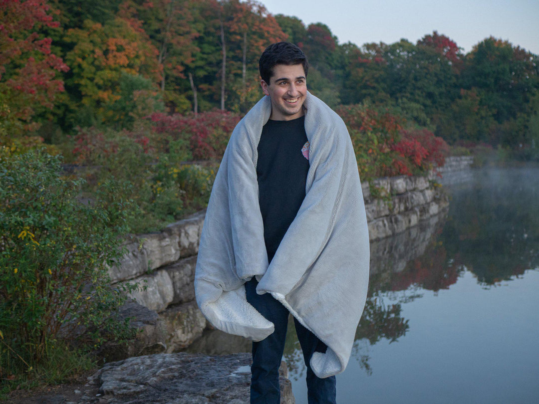 Man standing outdoors with black shirt and jeans wrapped in a Hush Grey Weighted throw blanket among red and green fall foliage and tress with rock face barrier bordering on a lake 