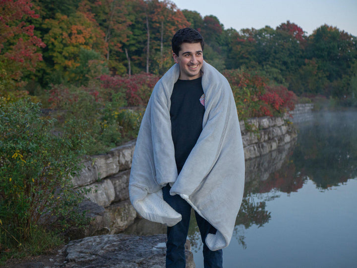 Man standing outdoors with black shirt and jeans wrapped in a Hush Grey Weighted throw blanket among red and green fall foliage and tress with rock face barrier bordering on a lake 