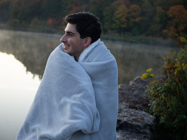 Man standing outdoors wrapped in a Grey Hush Weighted blanket with a lake and fall green and red trees in background