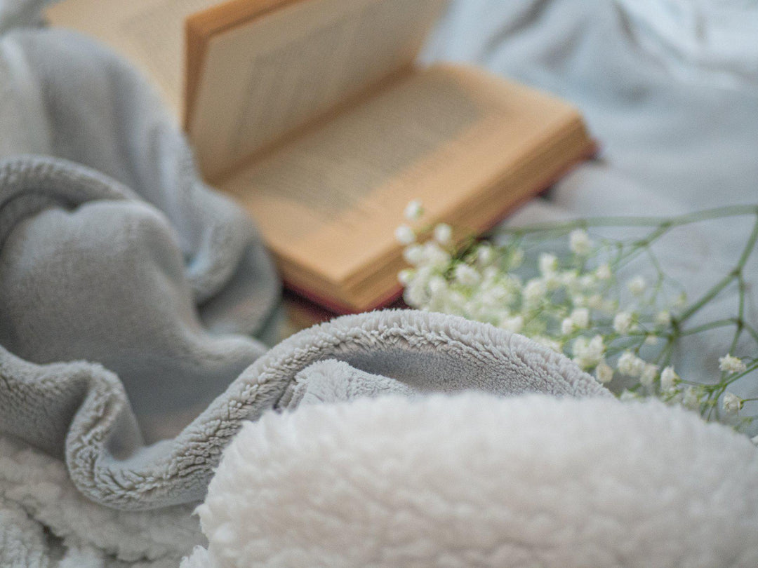 Hush weighted throw blanket in grey with white fleece liner and an open book and baby's breath flowers in background